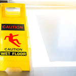 Gelbes Warnschild mit Aufschrift Caution Wet Floor warnt vor rutschigem Boden zur Vermeidung von Stolper- und Rutschunfällen.