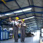 Zwei Beschäftigte mit Schutzhelm gehen durch eine Produktionshalle und besprechen Sicherheits- und Präventionsmaßnahmen.