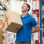 Mitarbeiter im Lager hebt schweren Karton falsch und fasst sich mit Schmerzen an den Rücken – typische Ursache für Rückenverletzungen durch falsches Heben.