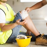 Verunfallter Bauarbeiter sitzt mit blutender, bandagierter Knieverletzung auf der Baustelle, Schutzhelm liegt neben ihm – Folge eines Stolper- oder Sturzunfalls.