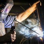 Schweißer mit Schutzschild arbeitet an Metallkonstruktion, Funken fliegen – Risiko für Verbrennungen und Augenverletzungen beim Schweißen.