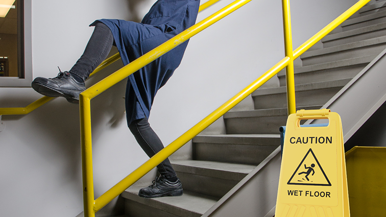 Darstellung einer Treppe, die eine potenzielle Gefahrensituation für einen Betriebsunfall darstellt. Mitarbeiter rutscht fast aus, kann sich aber noch am Geländer einhalten.
