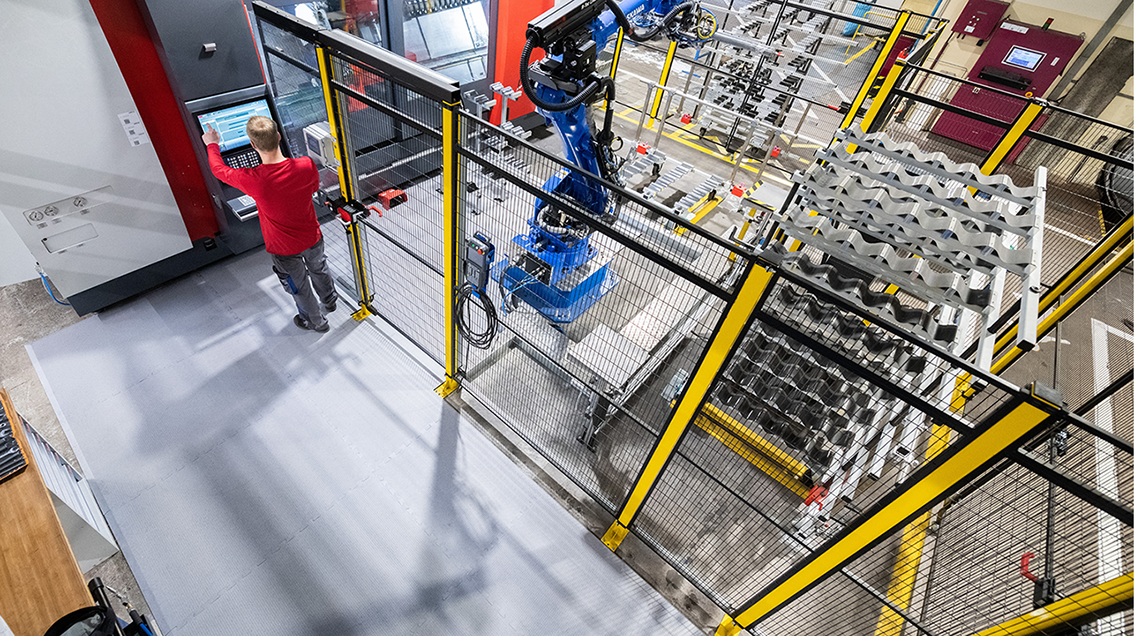 Vogelperspektive auf moderne Industriehalle: Bediener steuert Roboteranlage am Terminal, davor helle modulare KRAIBURG ERGOLASTECA Arbeitsplatzmatten als ergonomischer Boden – Industriearbeitsplatz erweitern mit flexiblem Bodensystem.