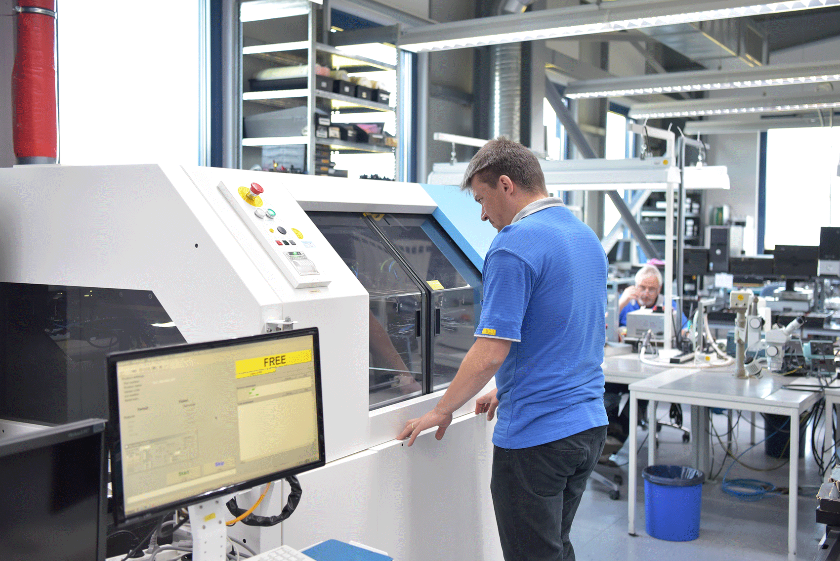 Ein Mann arbeitet an einer modernen Industrie- oder Produktionsmaschine in einem hellen, technisch ausgestatteten Labor oder Werkstattbereich. Im Vordergrund ist ein Computerbildschirm mit einer Steuerungssoftware zu sehen, im Hintergrund weitere Arbeitsplätze und technische Geräte.