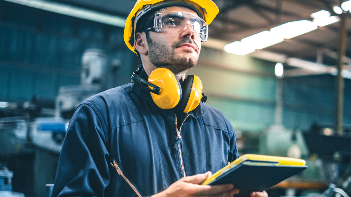 Industriemitarbeiter mit Schutzhelm, Gehörschutz und Tablet in der Hand blickt aufmerksam durch eine Produktionshalle – Symbol für gesetzeskonforme Ergonomie und Arbeitssicherheit am Arbeitsplatz.